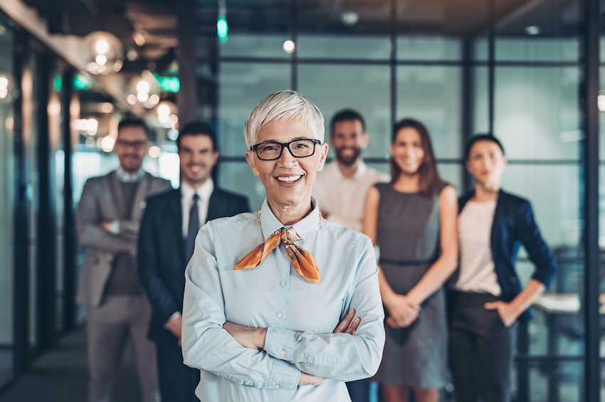Image of female professional standing with her arms-crossed, smiling widely in the foreground as a younger cohort of employees smile in the background representing the kind of joyful cohesive team that can exist when leadership communication is enhanced to foster trust and transparency 